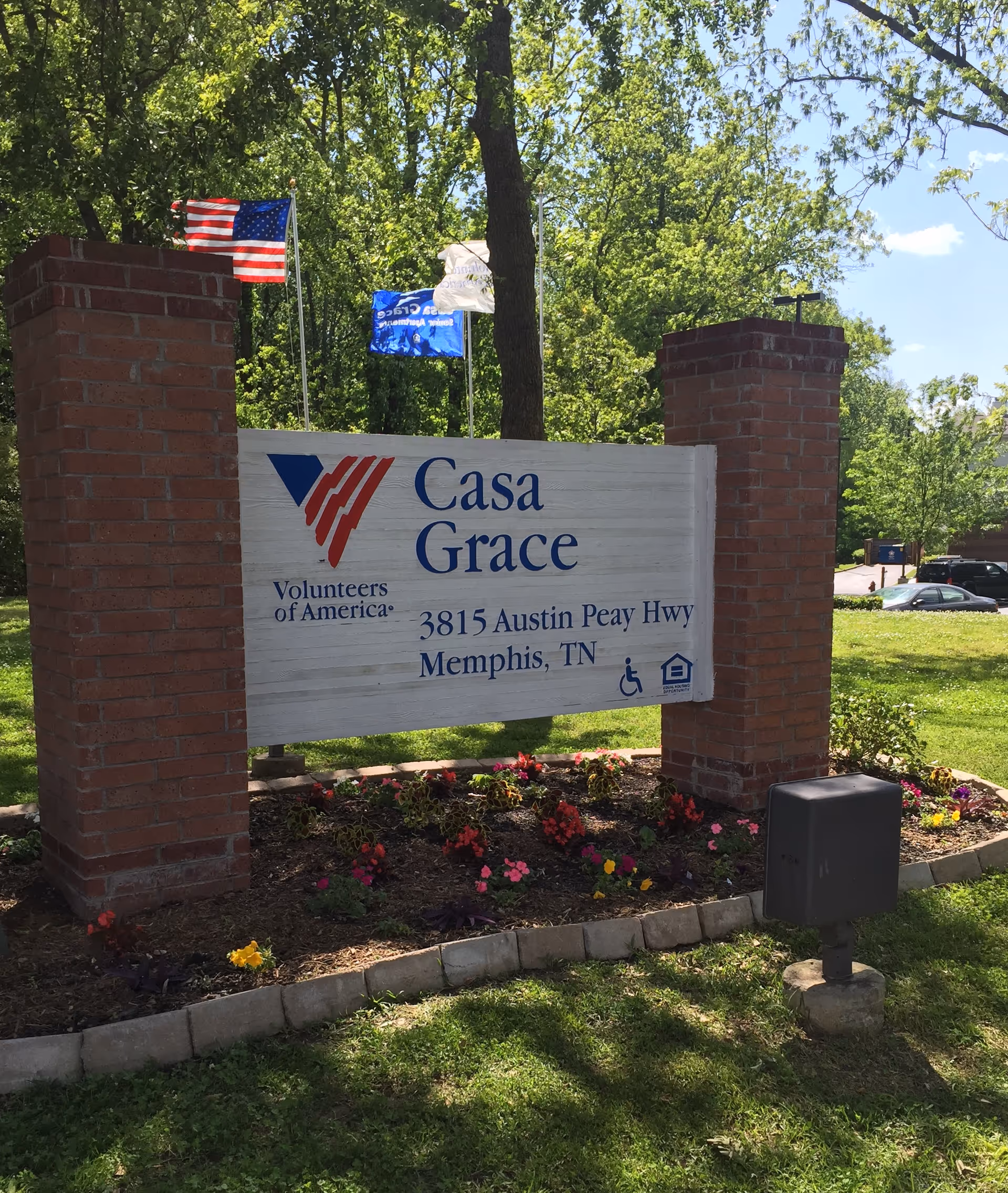 Entrance sign for Casa Grace Senior Housing with flags, brick pillars, and blooming flowers.