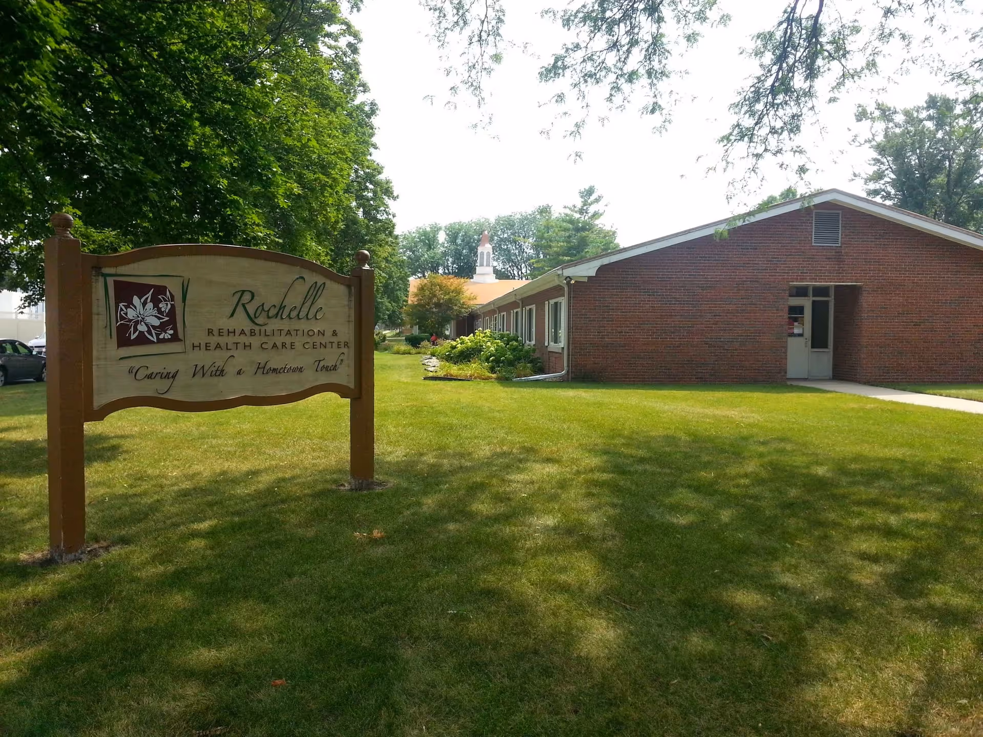 A wooden sign for 'Rochelle Rehabilitation & Health Care Center' stands on a grassy lawn in front of a single-story brick building and trees.