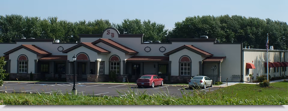 Front exterior of a single-story senior living building with arched windows, red awnings, and two cars in the parking lot.