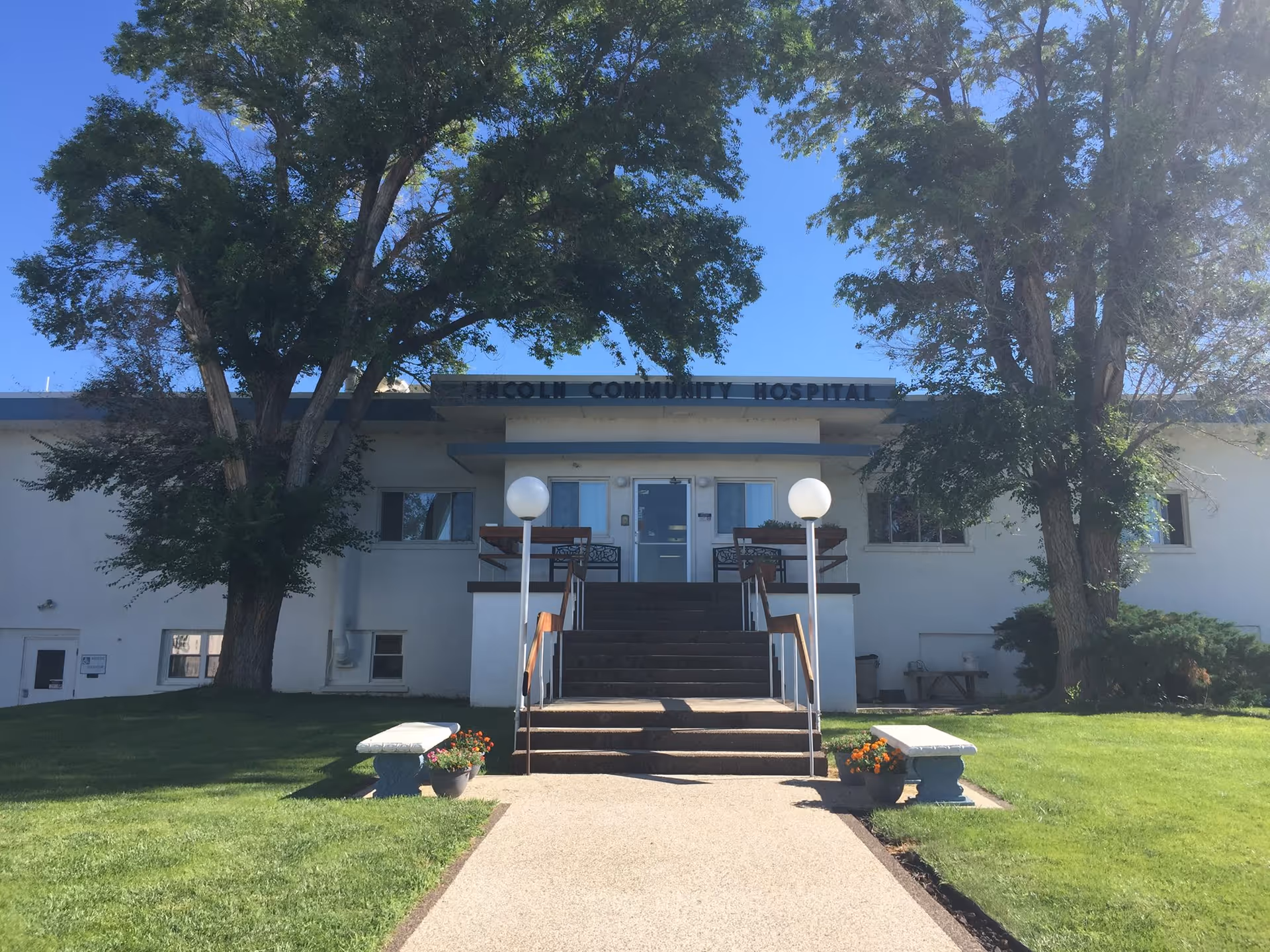 Front exterior of Lincoln Community Hospital with stairs, lamps, trees, and a green lawn.