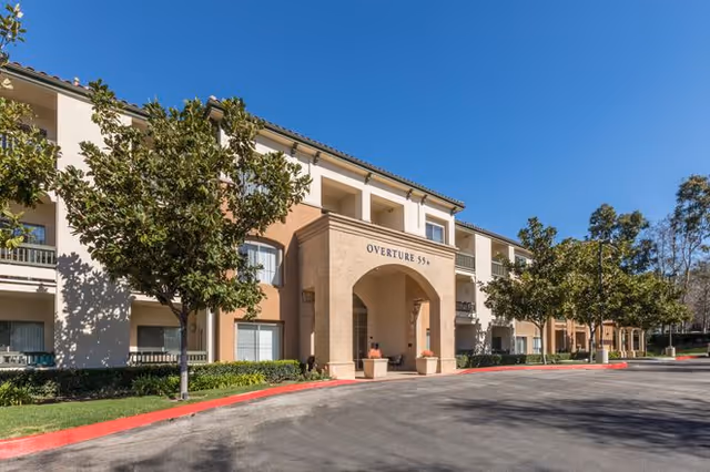 Front exterior of a three-story beige senior living building with an arched entrance labeled 'Overture 55+', trees, and a driveway under a clear blue sky.