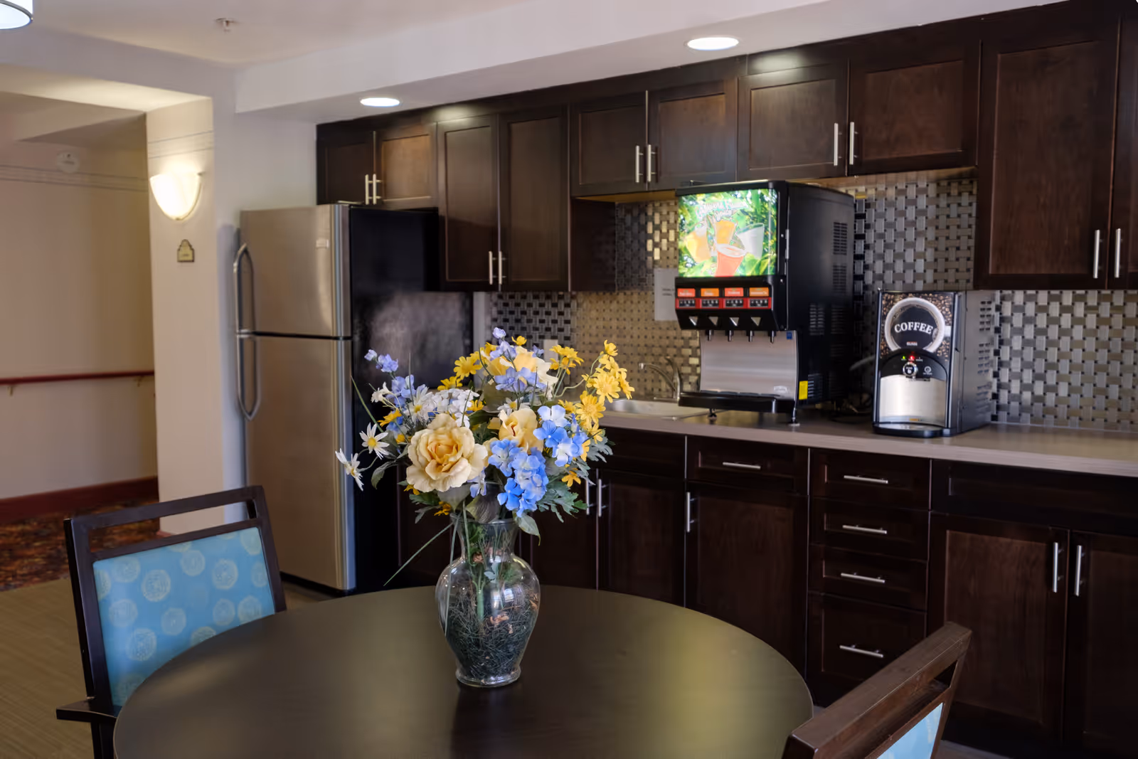 A kitchen area with dark wooden cabinets, a stainless steel refrigerator, a beverage dispenser, and a coffee machine on the counter. In the foreground, there is a round table with a vase of colorful artificial flowers, including yellow, blue, and white blooms. Two chairs with blue patterned cushions are partially visible around the table.