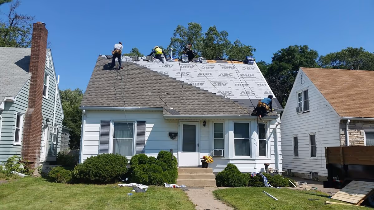 Several workers on a pitched roof installing shingles on a white house with a small front yard.