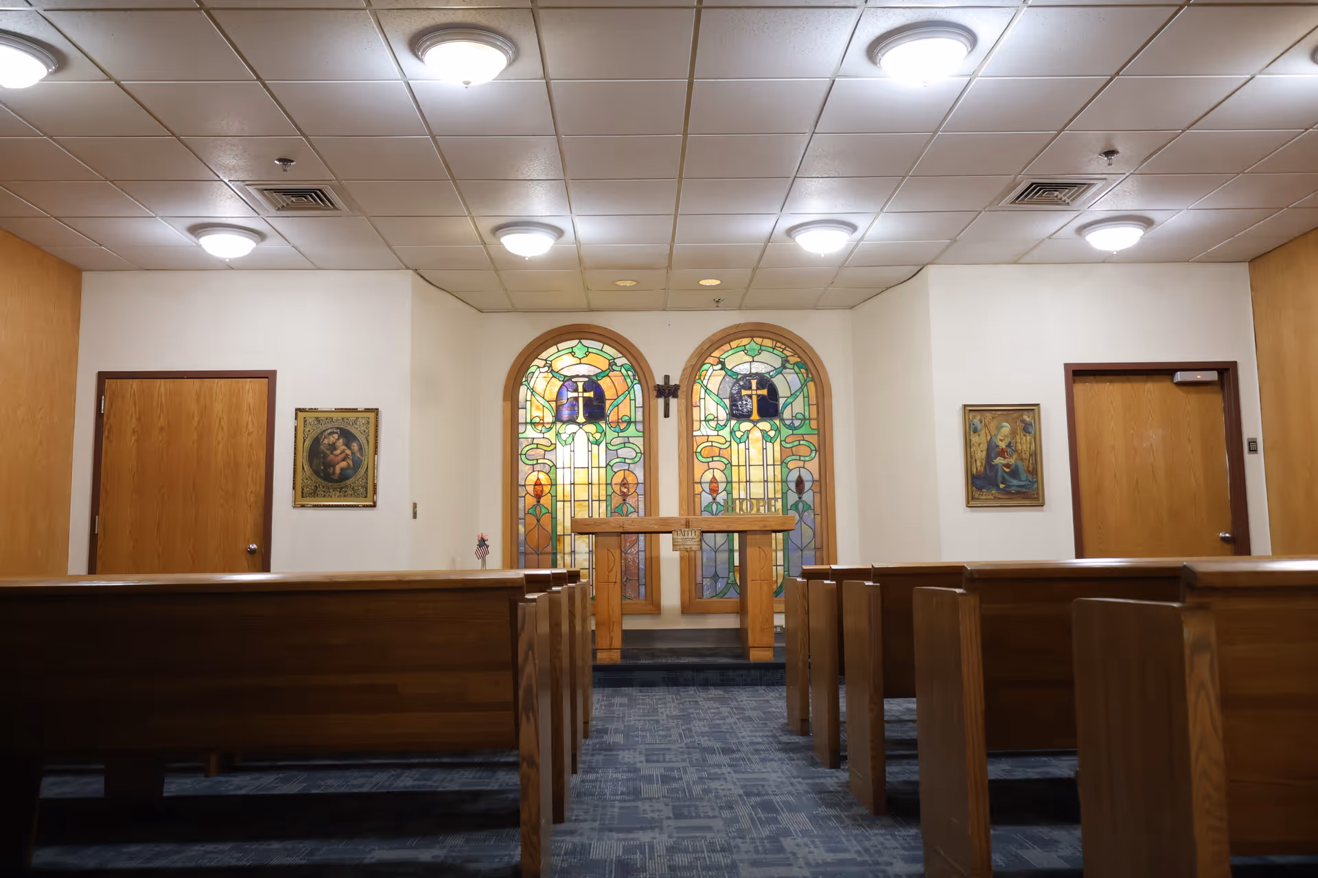 View of a chapel-like room with wooden pews, altar, and stained-glass windows.
