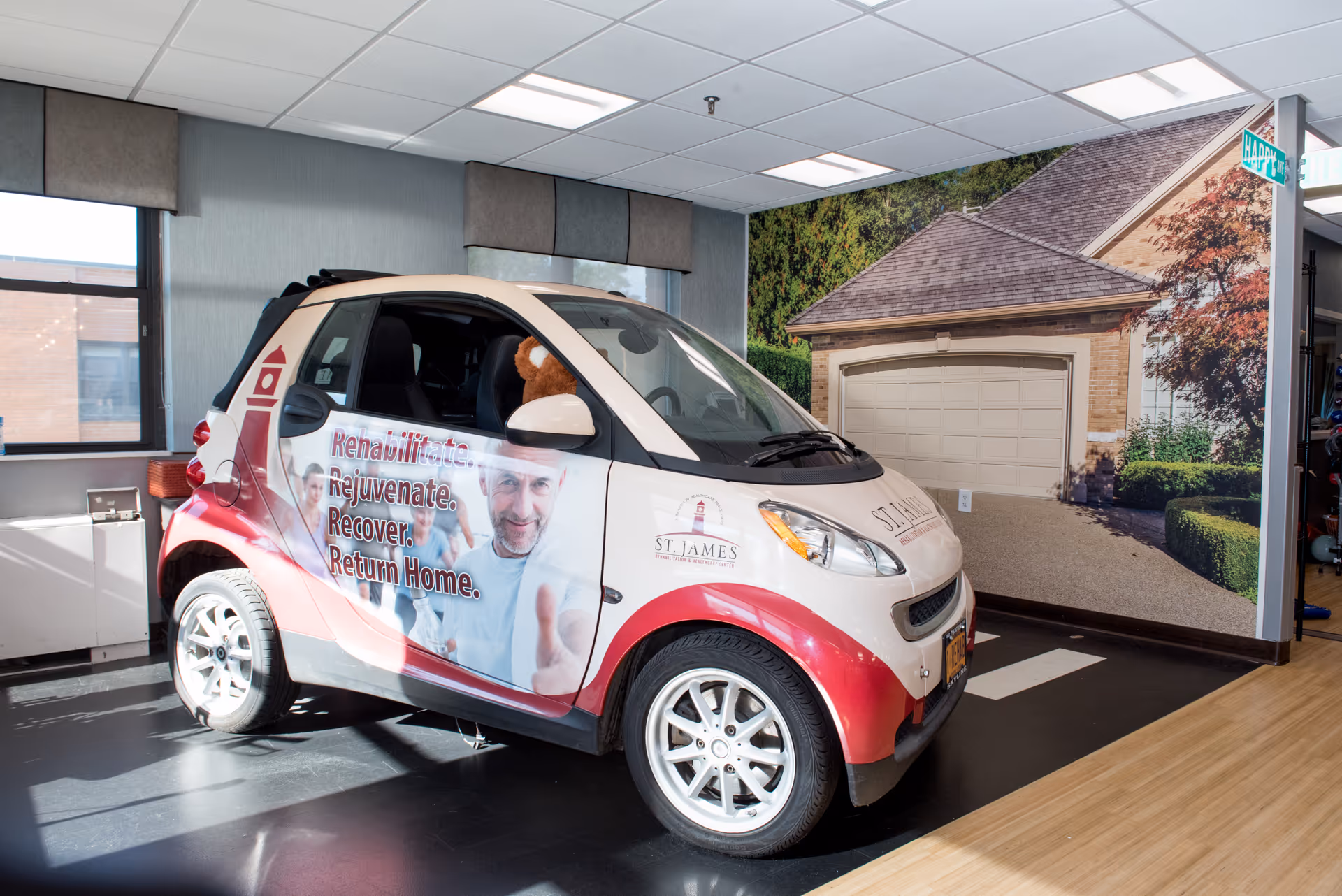 A small St. James–branded Smart car parked inside a facility lobby in front of a mural of a house garage.