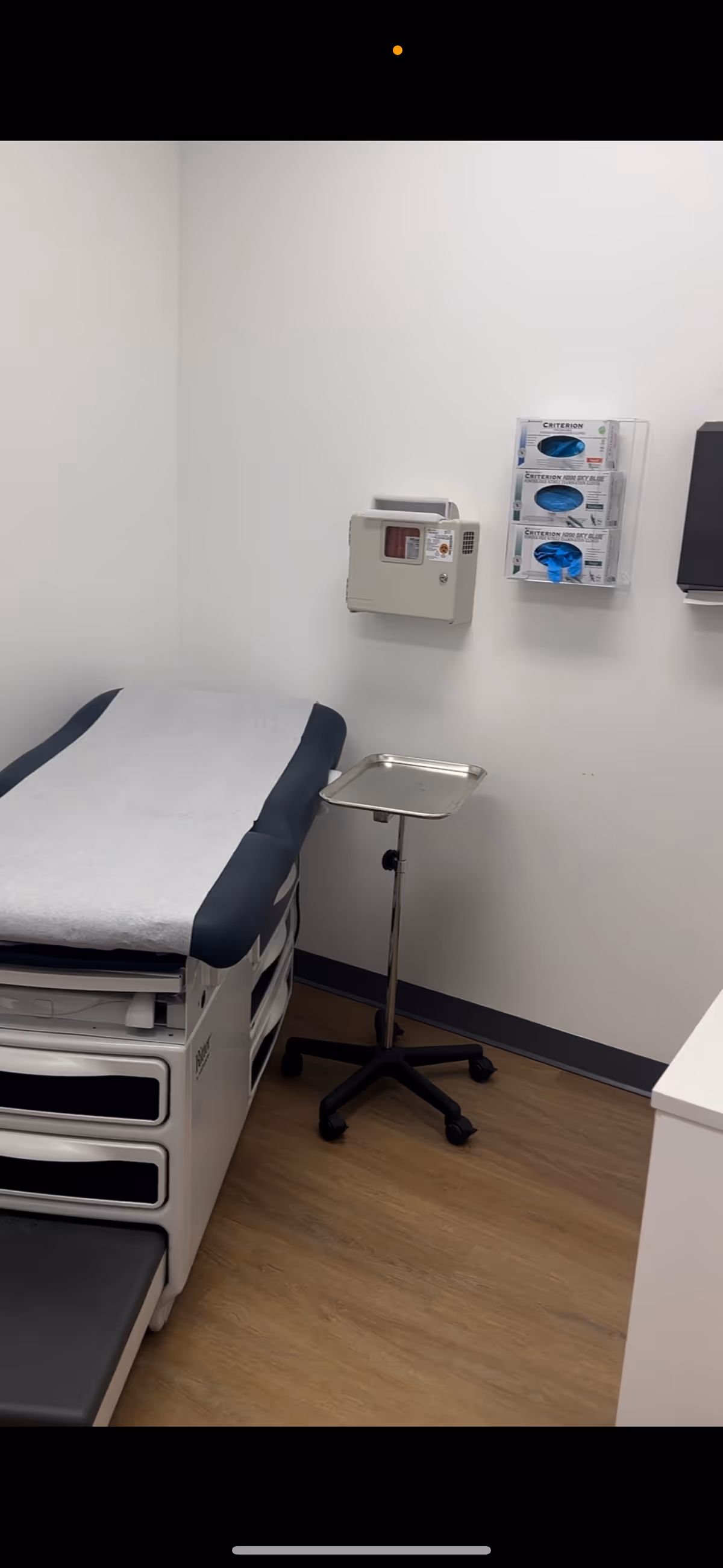 Interior clinical examination room with an adjustable bed, rolling stool, and wall-mounted supplies.
