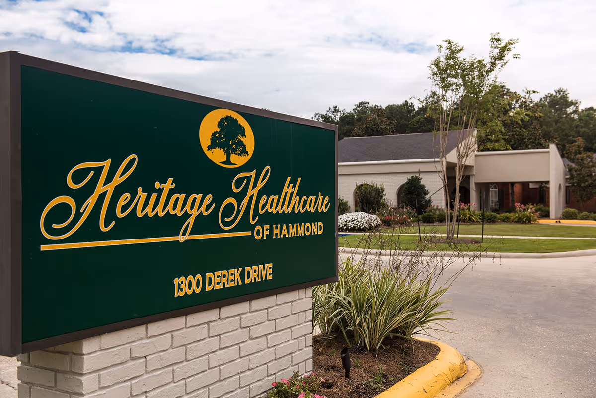 Outdoor view of Heritage Healthcare of Hammond sign with the address 1300 Derek Drive, situated on a brick base with landscaping around it. In the background, there is a single-story building with trees and shrubs under a cloudy sky.
