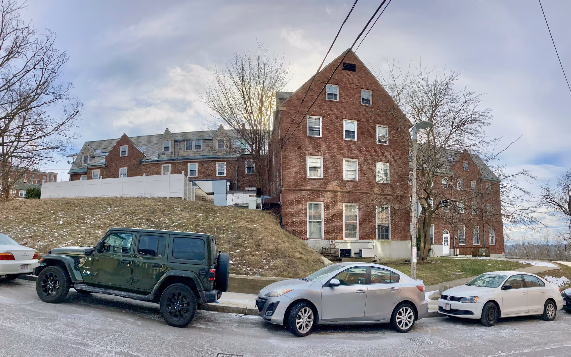 Exterior view of a large brick building with multiple windows and a steep roof, situated on a slightly elevated grassy area with some patches of snow. Several cars are parked along the street in front of the building, and leafless trees are visible around the property under a partly cloudy sky.