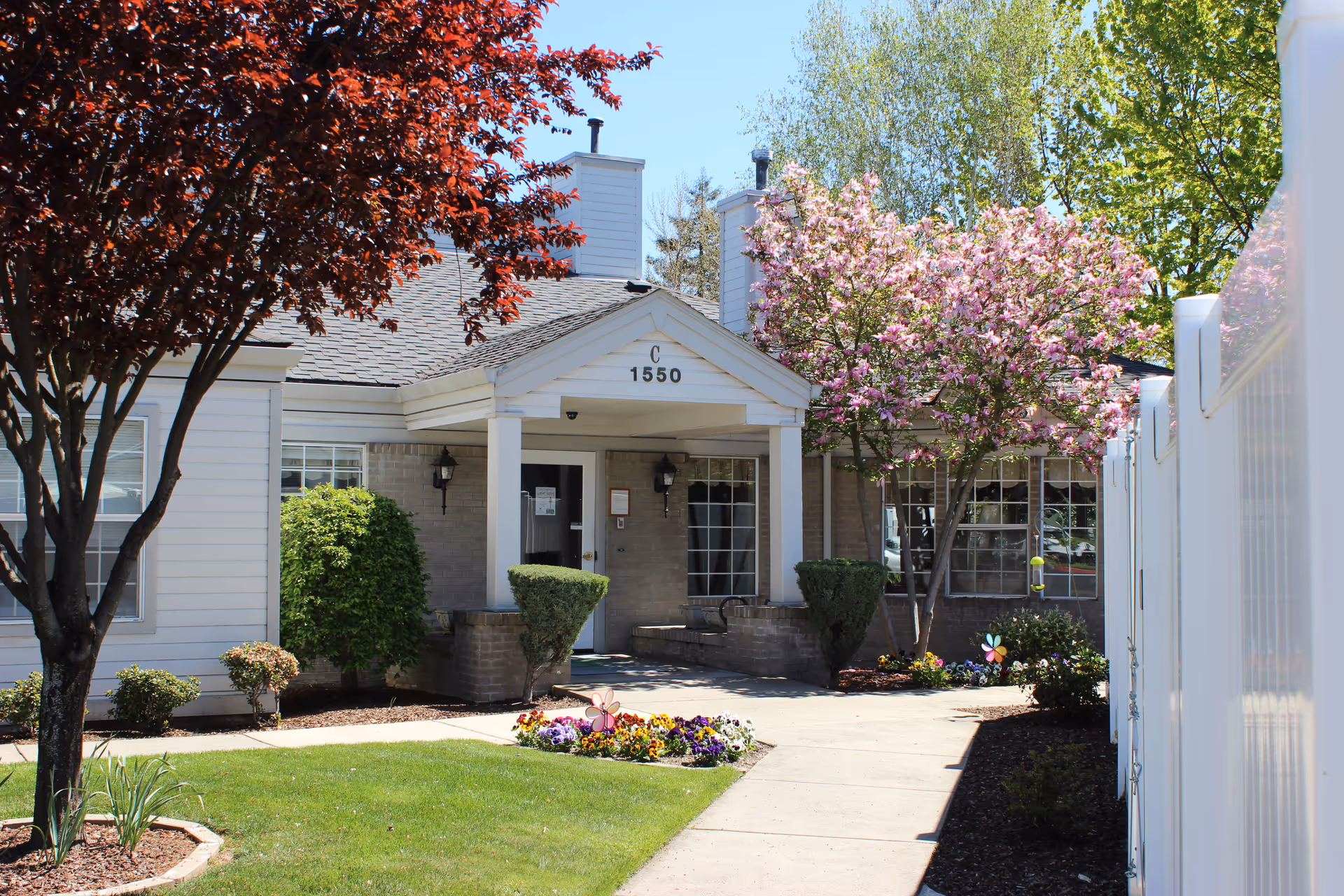 Front entrance of a low-rise building labeled 'C 1550' with a walkway, flowering trees, and manicured landscaping.