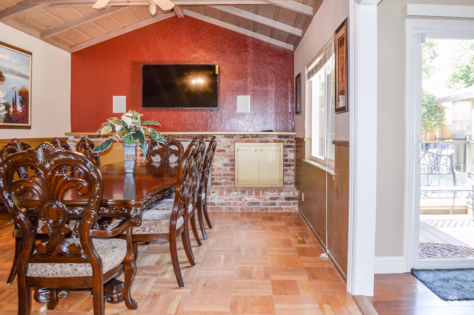 Dining room with a long wooden table surrounded by ornate wooden chairs with cushioned seats. A potted plant is placed in the center of the table. The room has a vaulted ceiling with exposed beams and a ceiling fan. A flat-screen TV is mounted on a red accent wall above a brick fireplace. Large windows and a glass door provide natural light and a view of an outdoor patio area.