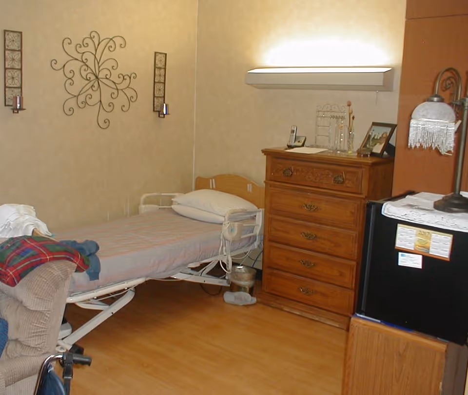 A small senior living bedroom with an adjustable hospital-style bed, wooden dresser, lamp and a small refrigerator.