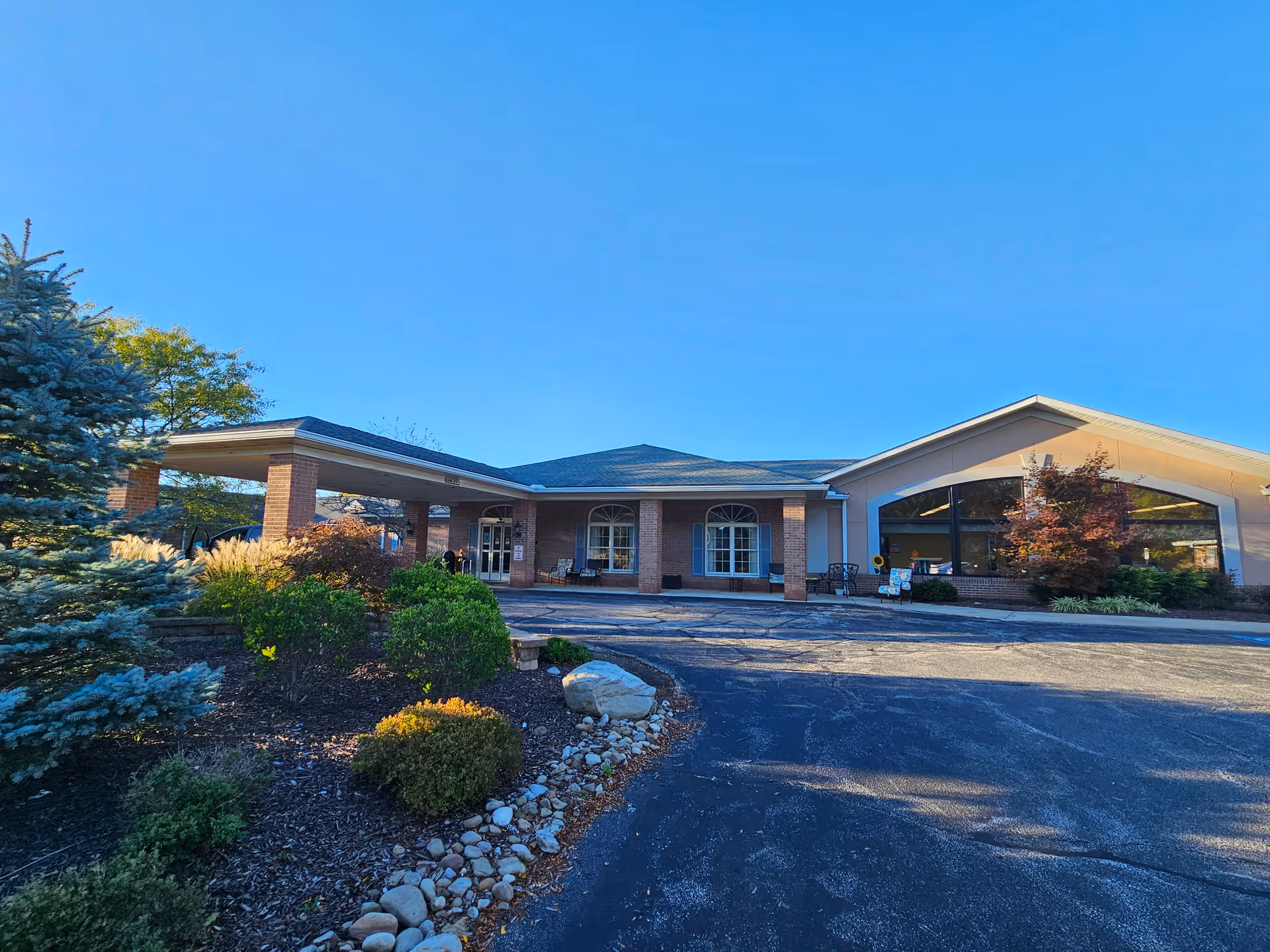 Front exterior of a single-story senior living facility with a covered entrance, large windows, and landscaped grounds under a clear blue sky.