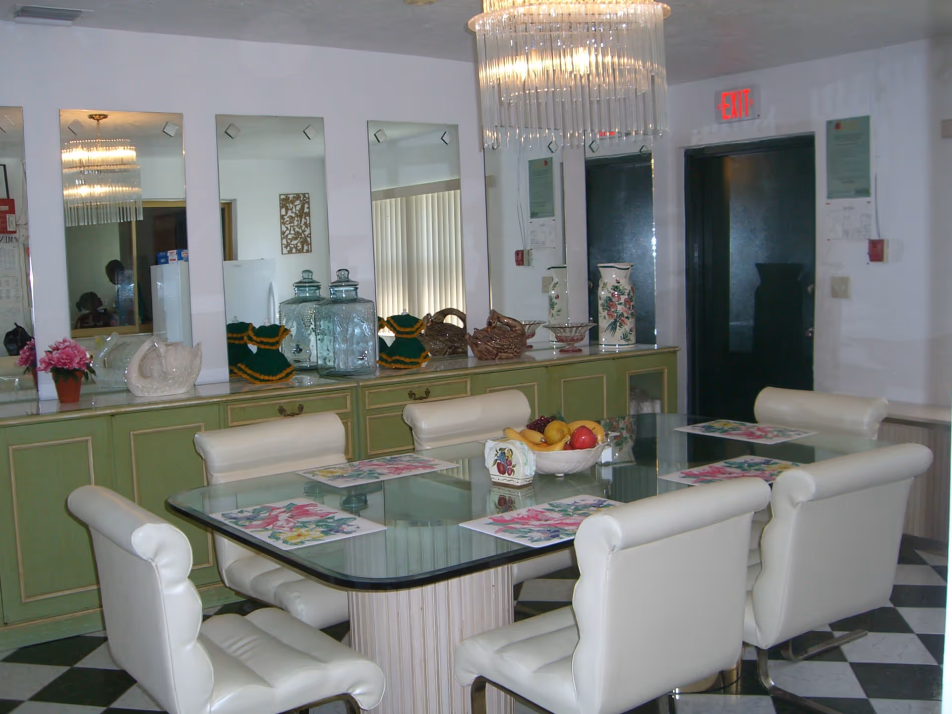 Dining room with a glass-top table surrounded by six white cushioned chairs. The table has floral placemats and a bowl of fruit in the center. Behind the table is a green sideboard with decorative items including glass jars, vases, and baskets. Four vertical mirrors hang above the sideboard, reflecting parts of the room. A chandelier hangs from the ceiling, and there is an exit door with a red exit sign above it.