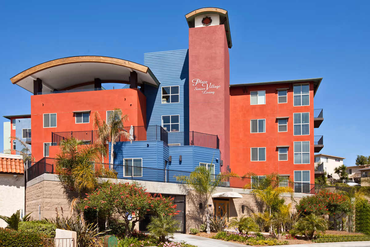 A colorful multi-story building labeled "Plaza Senior Village Living" with red and blue facades, balconies, palm trees, and a clear blue sky.