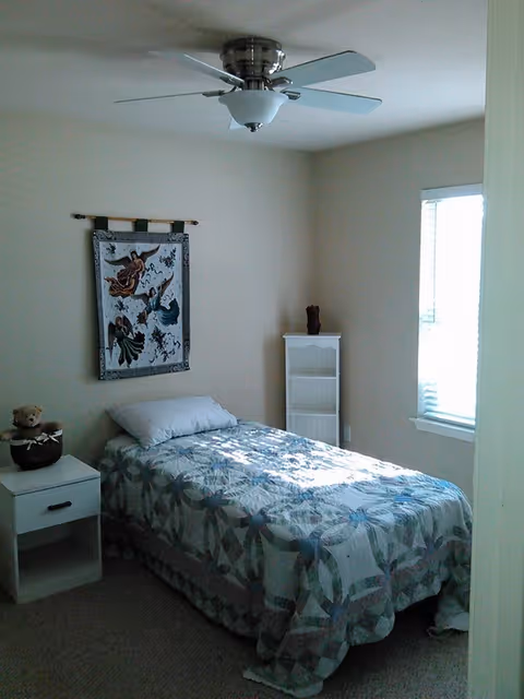 A small bedroom with a single bed, quilt, ceiling fan, and white nightstand.