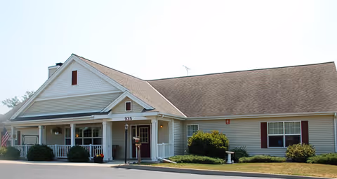 Exterior view of a single-story building with beige siding and a brown shingled roof. The building has a covered porch with white railings and columns, a lamp post near the entrance, and some bushes and landscaping in front. The address number 935 is visible above the porch.