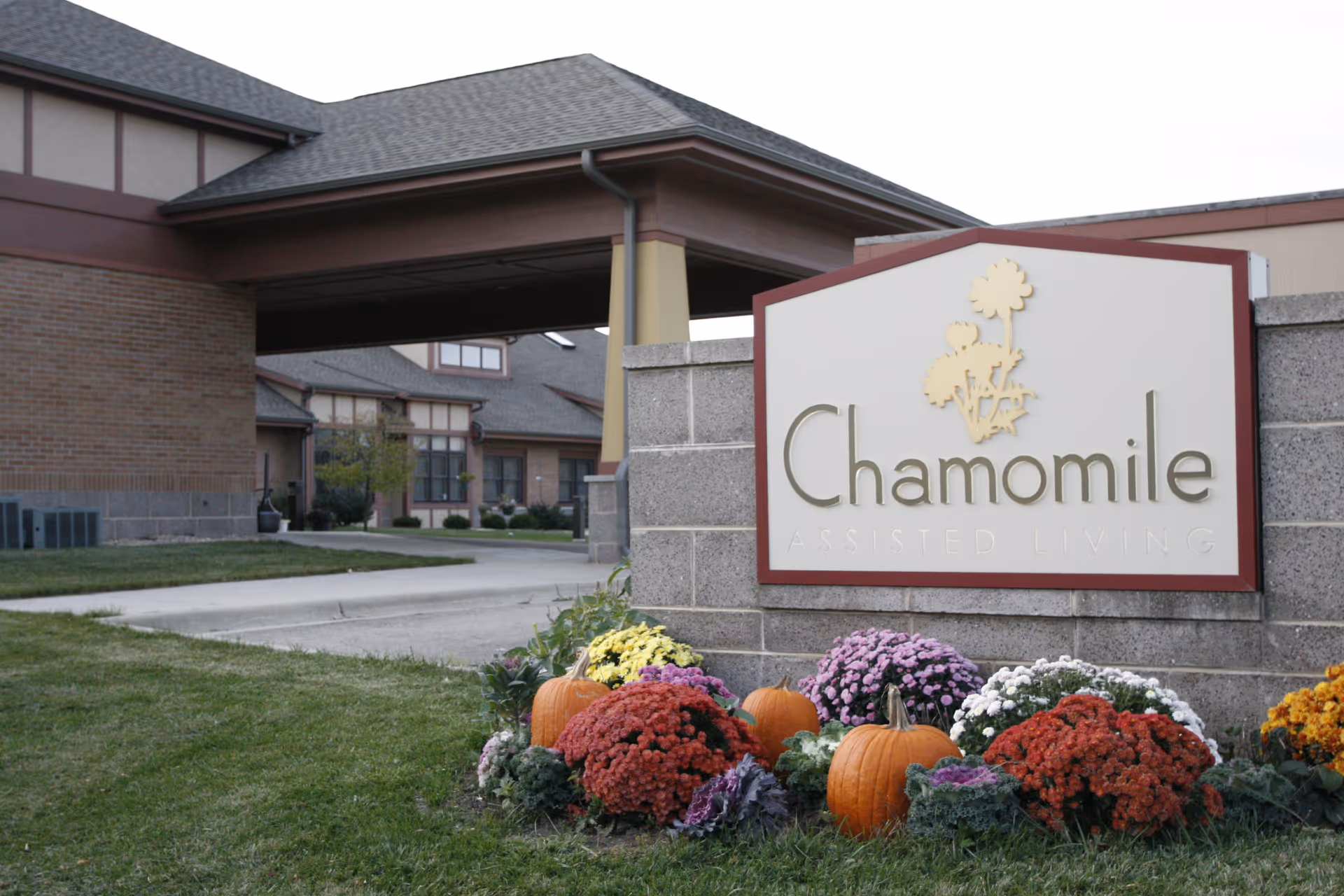 Entrance sign for Chamomile Assisted Living with flowers and pumpkins planted around the base, and the facility building visible in the background.
