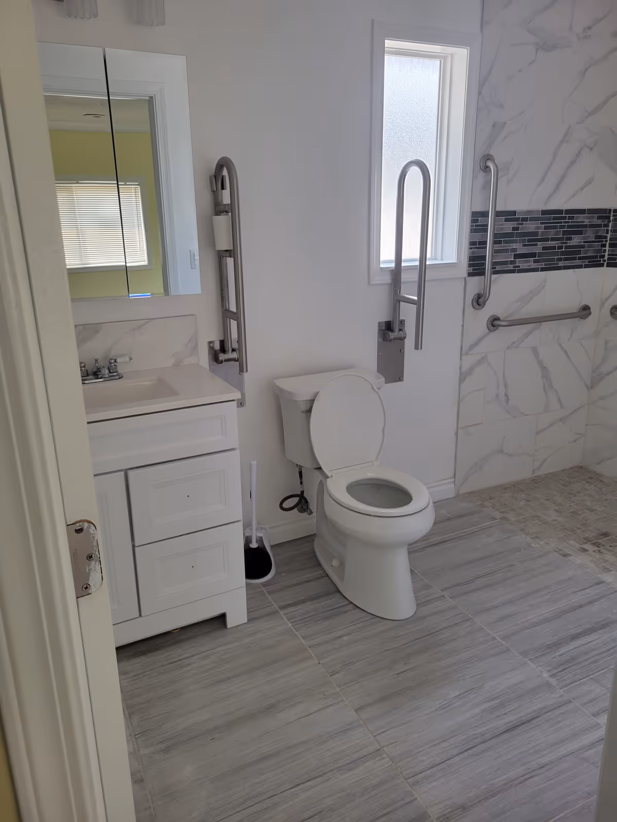A clean, accessible bathroom with a white toilet, white vanity with sink, and a mirror above. There are stainless steel grab bars around the toilet and in the shower area, which has a tiled wall with a decorative horizontal strip. The floor is tiled with large gray tiles, and there is a frosted window letting in natural light.
