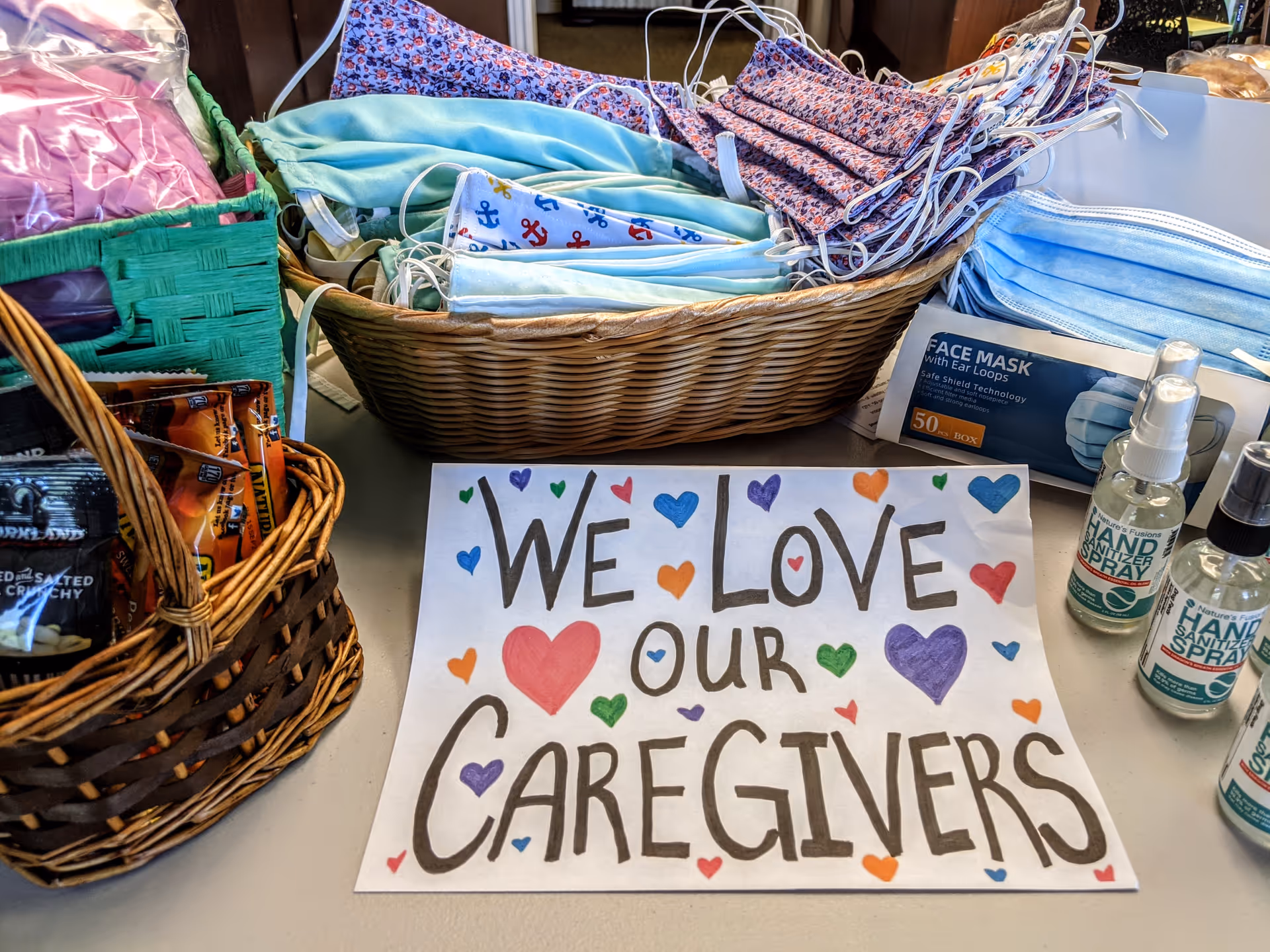A table displays baskets of masks and hand sanitizer with a handwritten sign thanking caregivers.