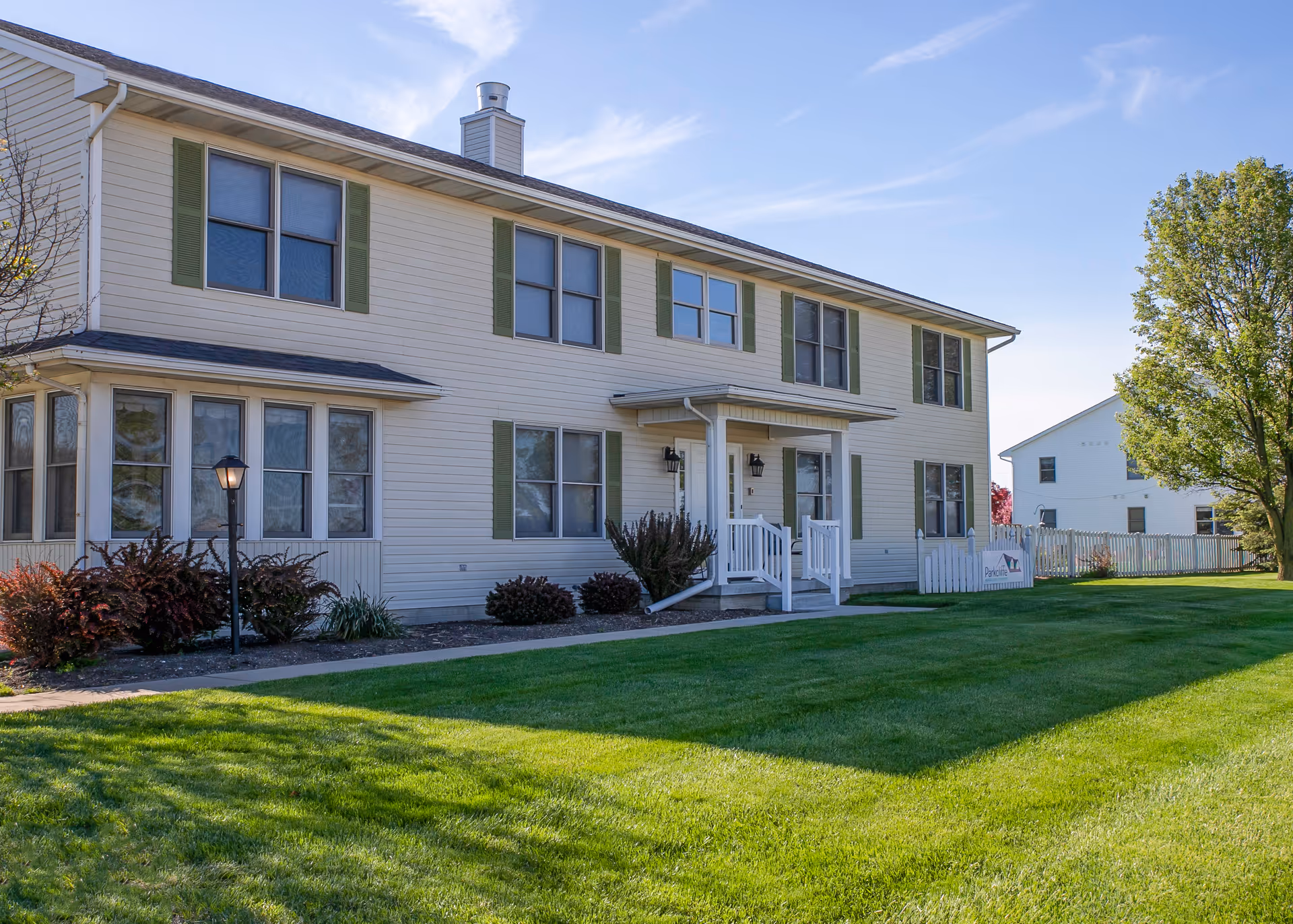 Two-story beige senior living facility with green shutters, front porch, and a well-kept lawn.