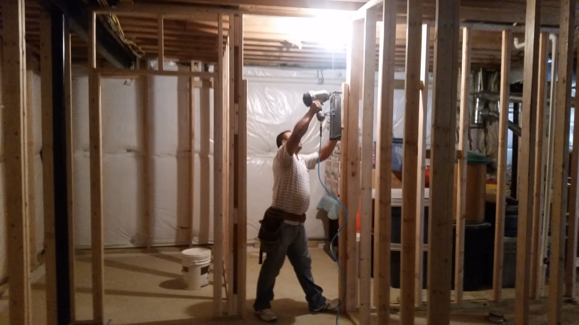 Man using a power drill inside an unfinished building with exposed studs.