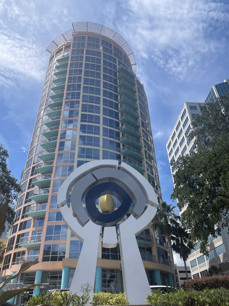 Tall curved glass high-rise with balconies and a white abstract sculpture in foreground.
