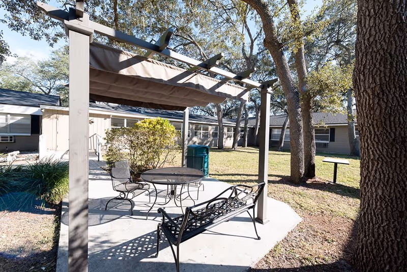 Shaded outdoor patio with a pergola, metal table and chairs and a bench in a grassy courtyard in front of a low building.