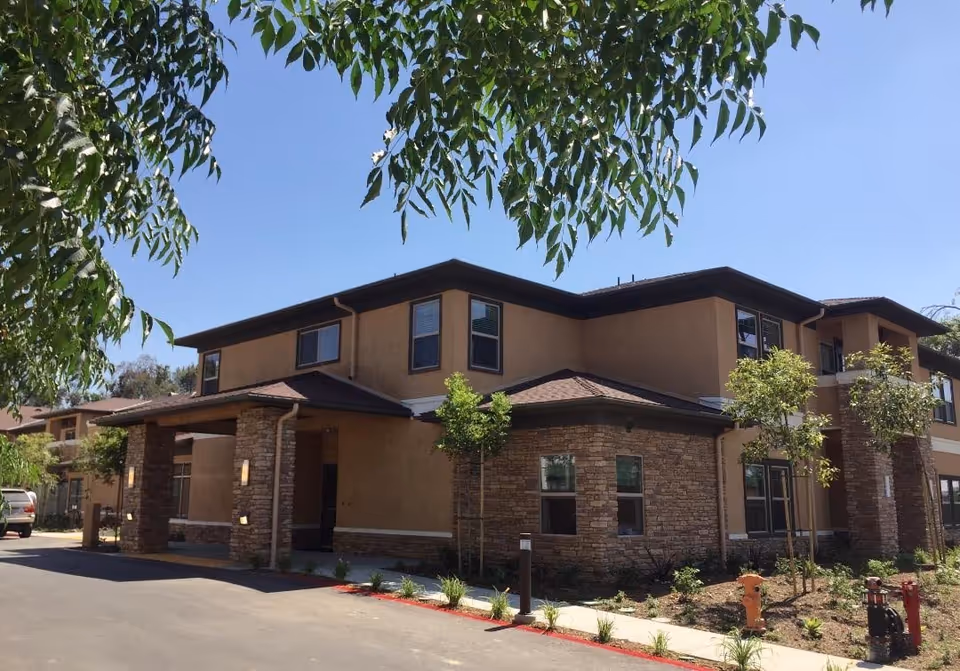 Two-story senior living building with stone accents, trees, and a driveway in the foreground.