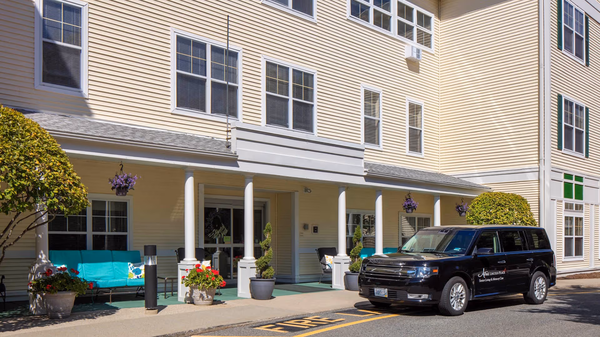 Exterior view of Atria Lincoln Place senior living facility showing the entrance with white columns, potted plants, and a black vehicle parked in front with the facility's logo on the side.