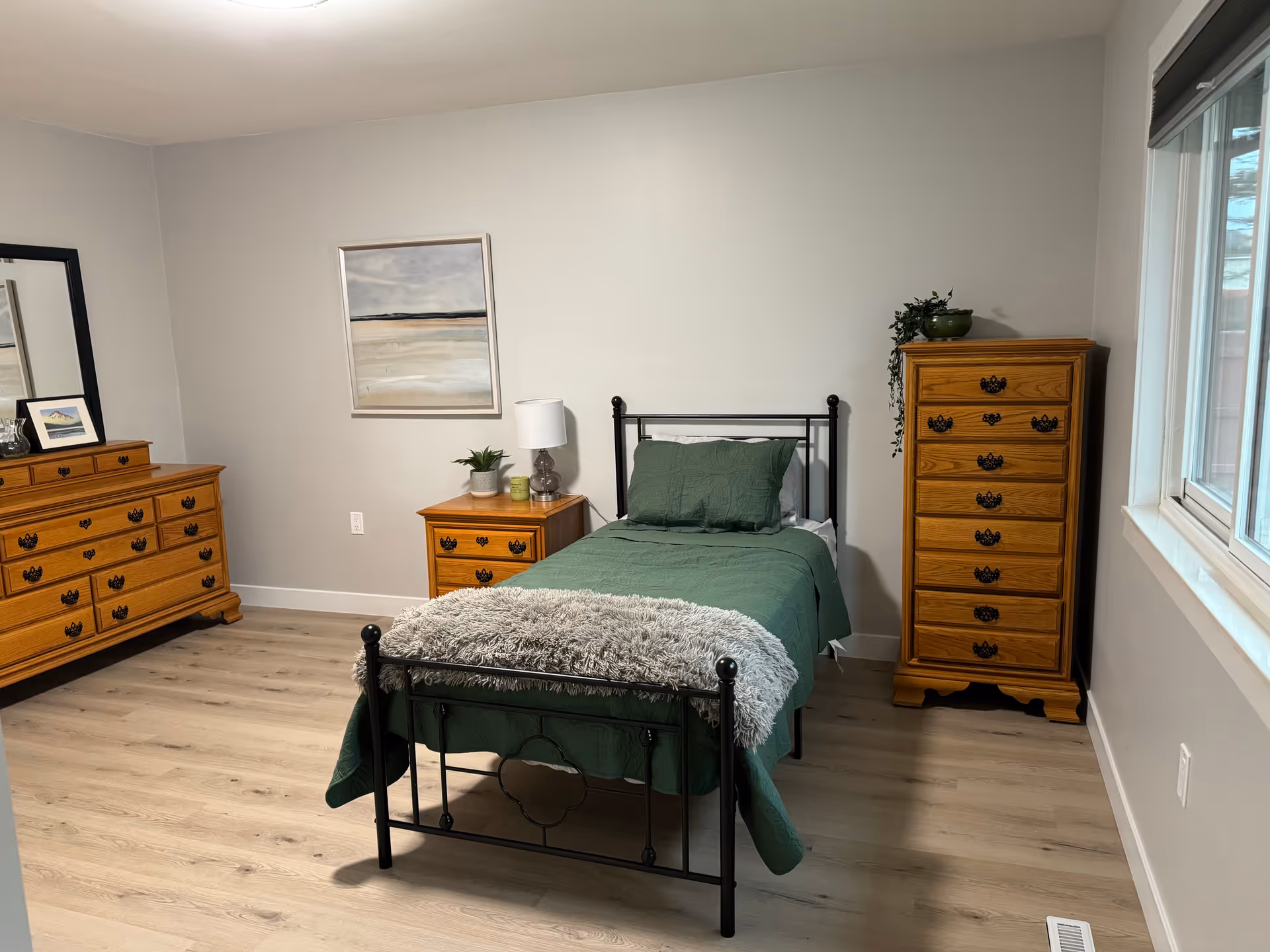 A furnished Trivol Manor bedroom with a metal-framed bed, green bedding, and wooden dressers.