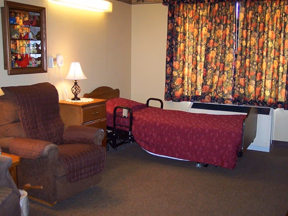 A cozy retirement facility bedroom with a single bed covered in a red patterned bedspread, a brown recliner chair with a quilted cover, a wooden nightstand with a lamp, and floral curtains covering the window.