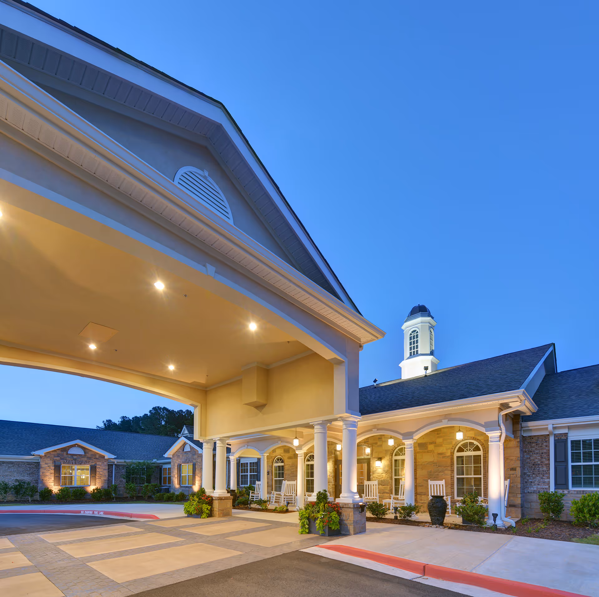Exterior view of Benton House of Woodstock senior living facility at dusk, featuring a covered entrance with white columns, rocking chairs on the porch, and warm lighting illuminating the building's stone and brick facade.
