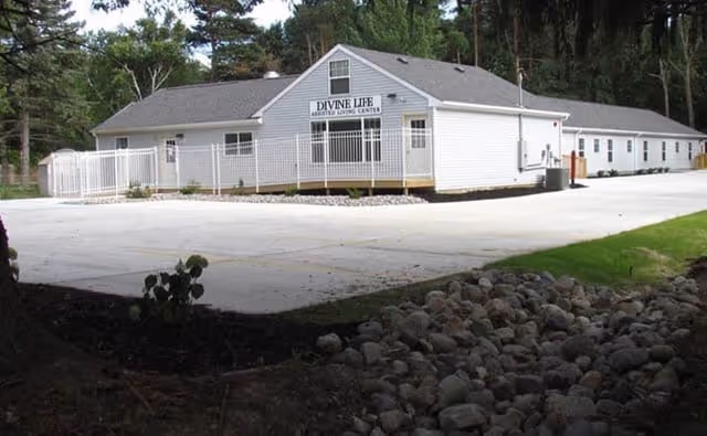 Single-story white assisted living building with a sign reading 'Divine Life Assisted Living Center', a fenced entrance, and a paved driveway surrounded by trees.