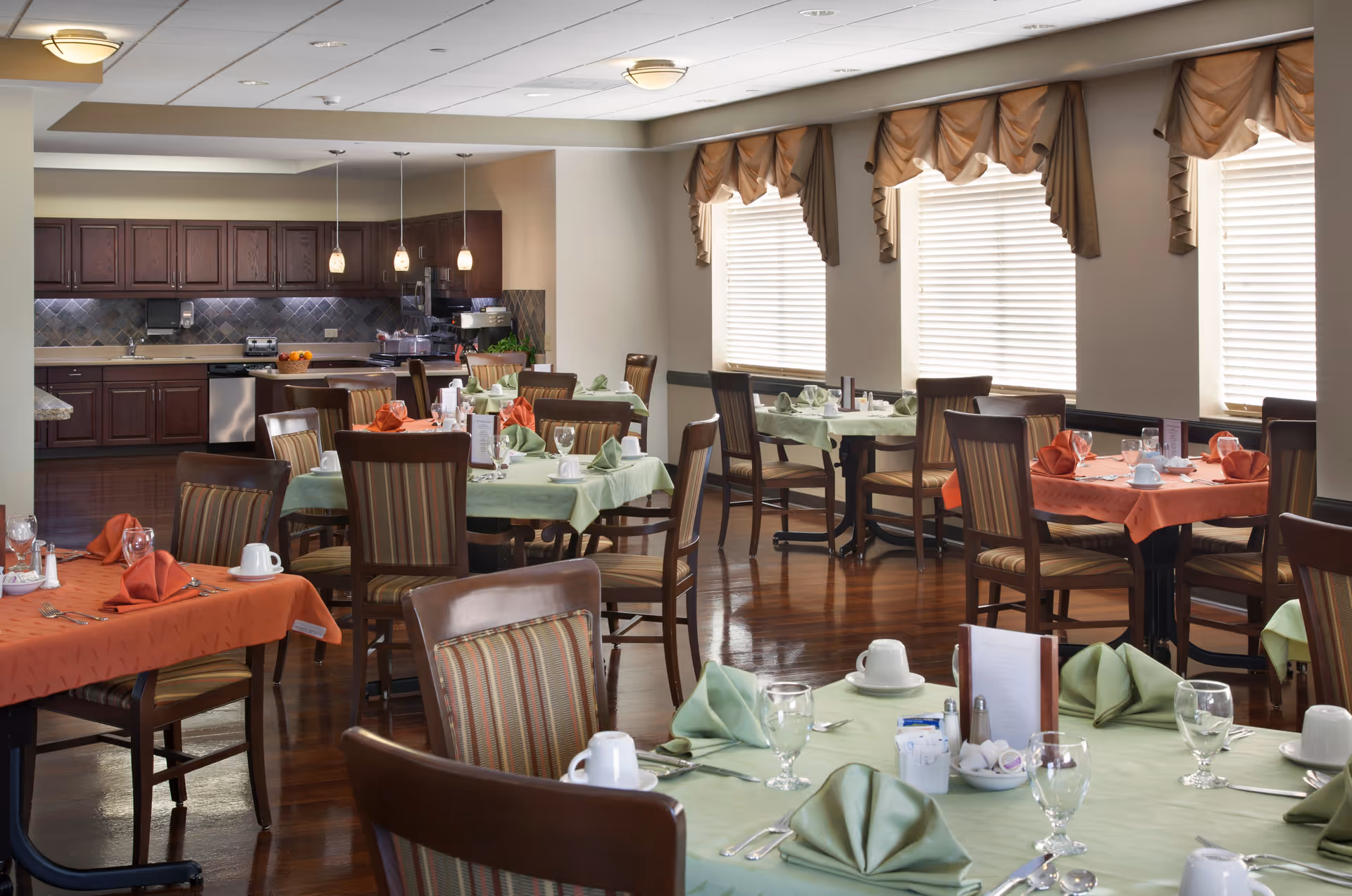 A dining room in a senior living facility with multiple tables covered in green and orange tablecloths, each set with folded napkins, glasses, cups, and silverware. The room has wooden chairs with striped cushions, large windows with blinds and valances, and a kitchen area with dark wood cabinets and stainless steel appliances in the background.