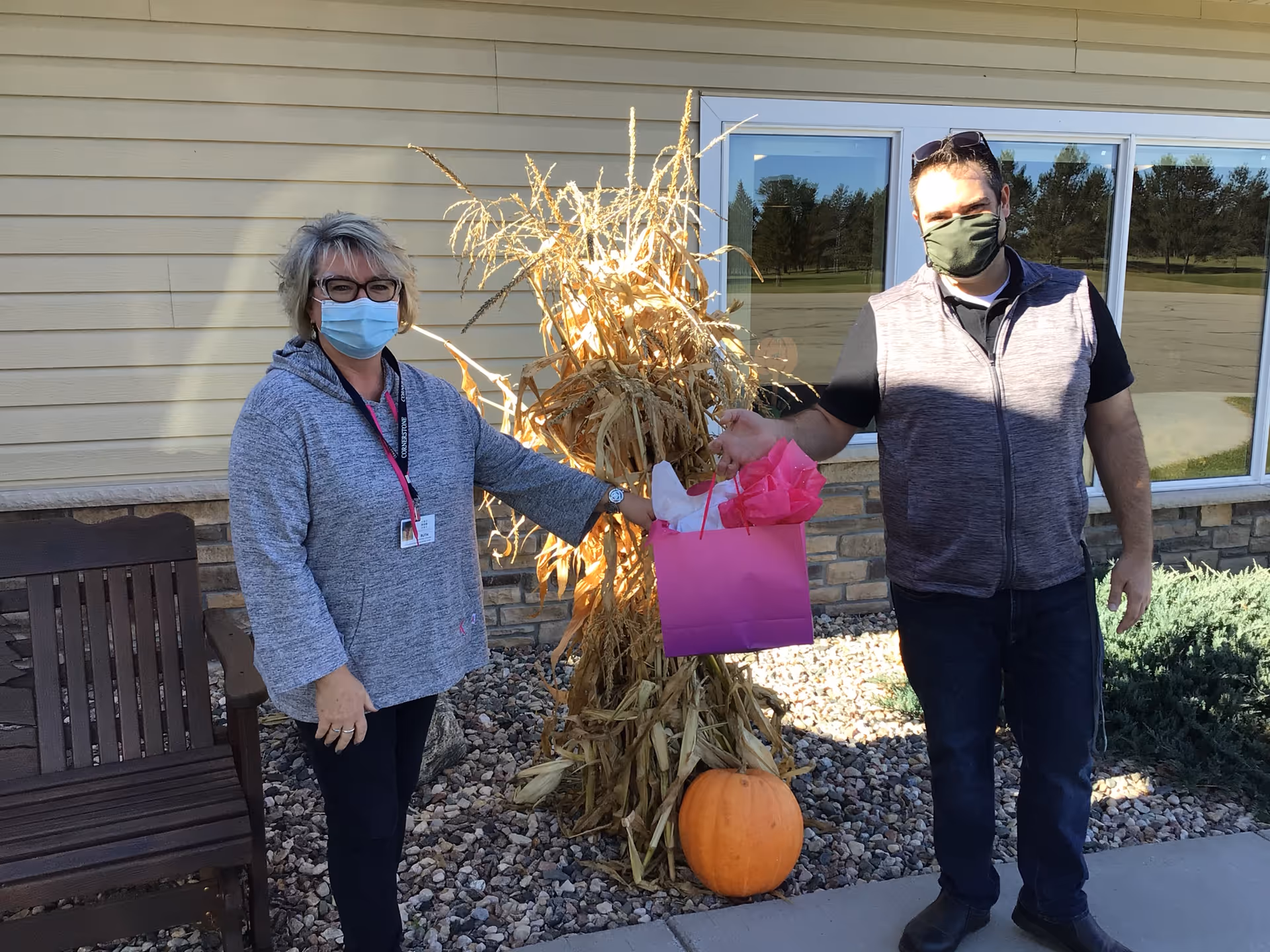 Two people wearing face masks stand outside a building next to a decorative bundle of dried corn stalks and a pumpkin. One person is holding a pink gift bag with tissue paper. There is a wooden bench to the left and windows reflecting trees in the background.