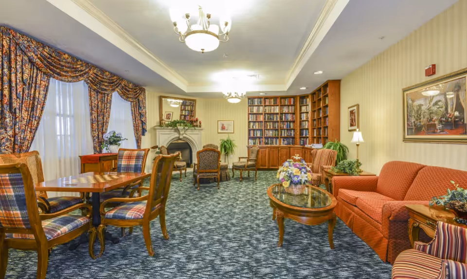 A cozy living room area with patterned carpet, a red sofa, striped armchair, wooden coffee table with a flower arrangement, and a wooden dining table with plaid upholstered chairs. The room features large windows with floral curtains, a fireplace with a mirror above it, built-in bookshelves filled with books, and framed artwork on the walls.