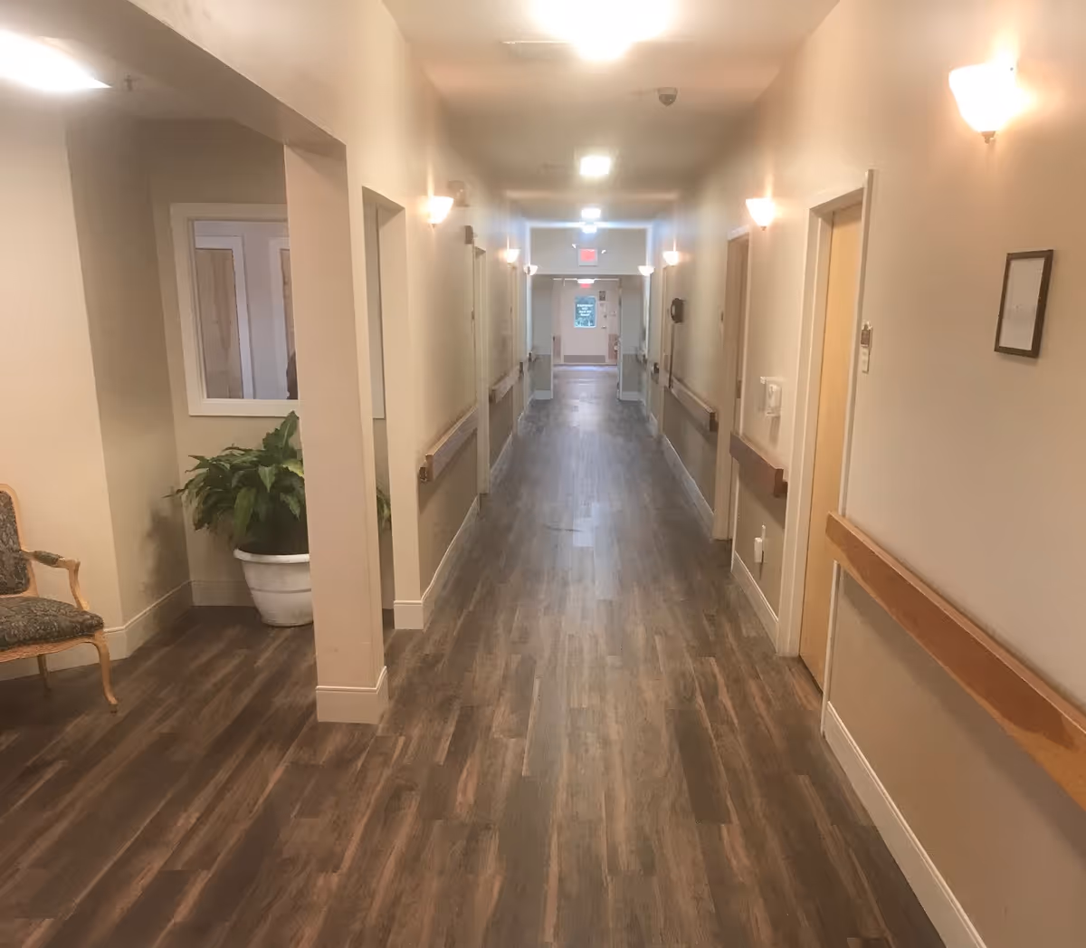 Long indoor hallway in a senior living facility with a plant and a chair.