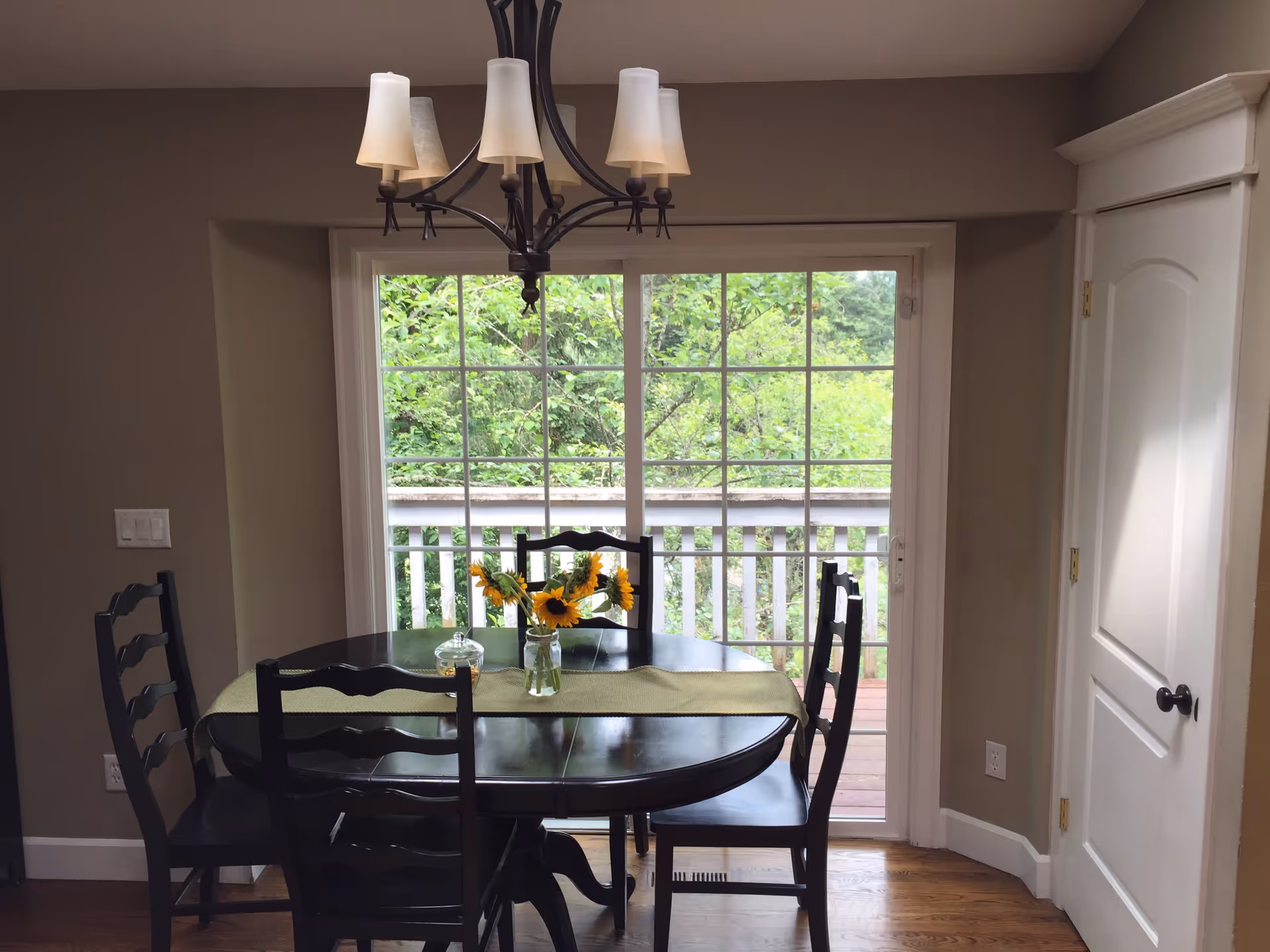 Dining area with a dark wood table and chairs, a vase of sunflowers, chandelier, and sliding glass door opening to a deck and greenery.