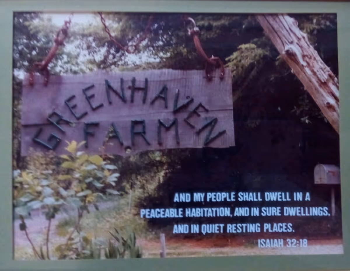Weathered wooden sign hangs among trees, with large carved letters and Isaiah 32:18 verse.