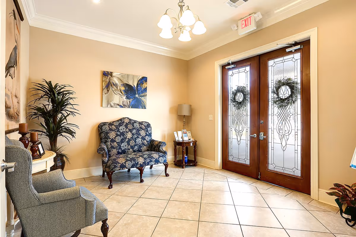 A small seating area near a set of double wooden doors with decorative glass panels and wreaths. The room has beige walls, tiled floor, a blue floral upholstered loveseat, a gray armchair, a small round wooden table with a lamp and brochures, a potted plant, and a painting of a blue butterfly on the wall.