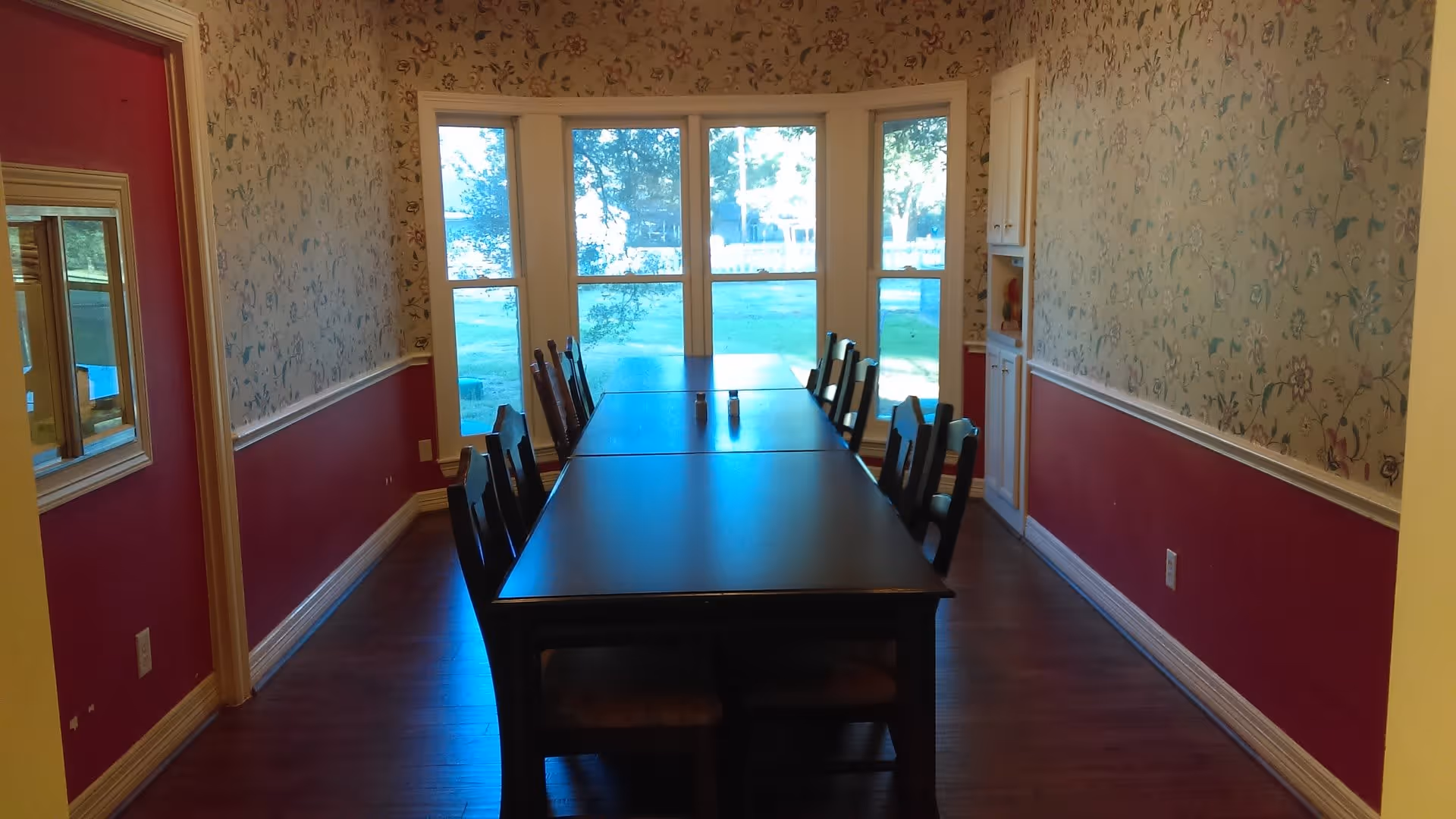 A dining room with a long rectangular dark wooden table surrounded by eight chairs. The walls have floral wallpaper on the upper half and red paint on the lower half. There is a large bay window at the far end letting in natural light and providing a view of green grass and trees outside. A built-in cabinet is visible on the right side of the room.