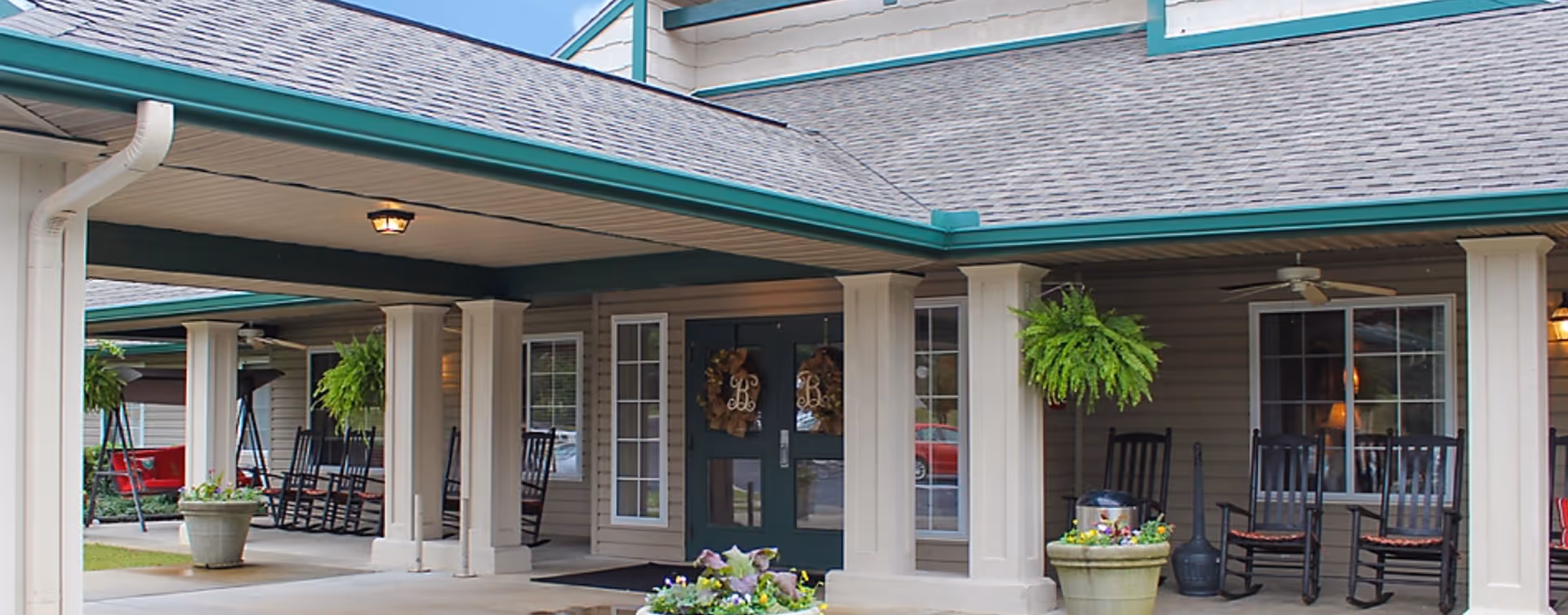 Exterior view of a senior living facility entrance with a covered porch, rocking chairs, and planters.