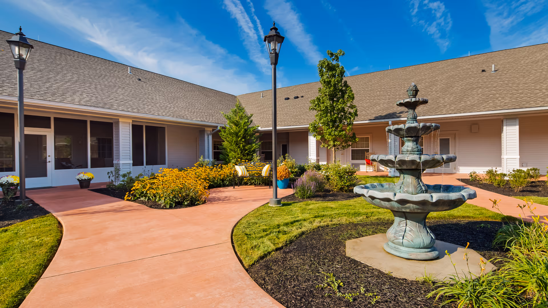 Outdoor courtyard area at Legend of Lititz featuring a three-tiered water fountain, curved walkways, green grass, flower beds with yellow flowers, small trees, benches, and a building with a covered porch under a blue sky with wispy clouds.