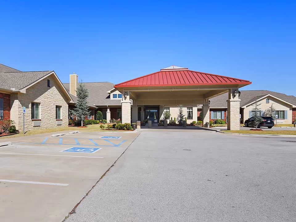 Front exterior view of a senior living facility with a covered entrance featuring a red roof, stone pillars, and a driveway with handicap parking spaces. The building has a combination of brick and stone walls under a clear blue sky.