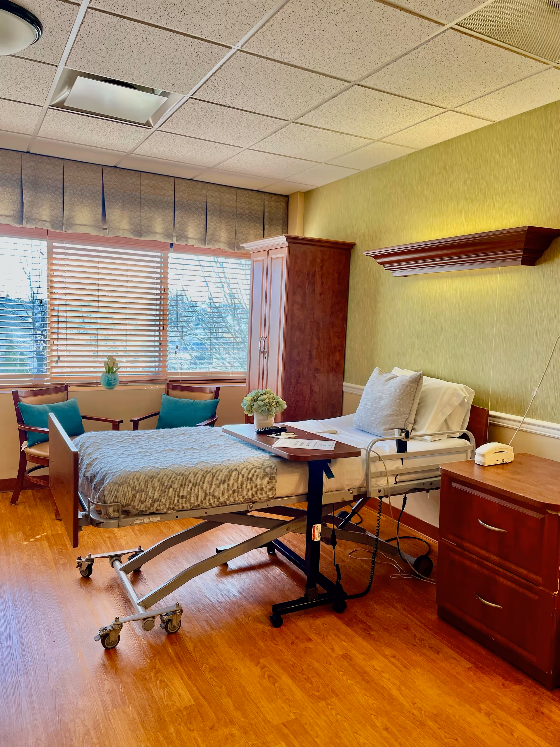 Hospital-style patient bed in a furnished room with window, dresser, and nightstand.