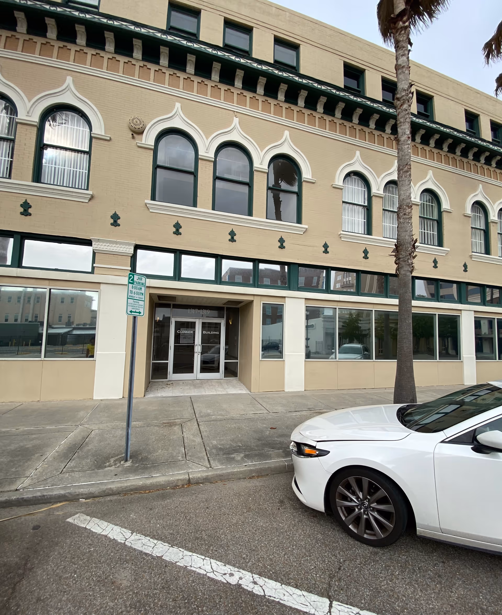 Exterior view of a brick building with arched windows, palm tree, and a parked white car.