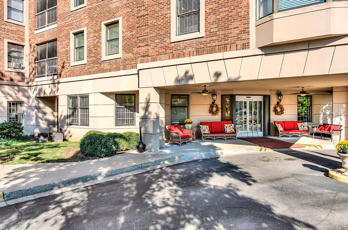 Entrance area of a brick and beige building with a covered porch featuring red cushioned wicker chairs and sofas, ceiling fans, decorative wreaths on the walls, and a glass automatic door. There are bushes and a small grassy area to the left, and shadows of trees are cast on the pavement and building.