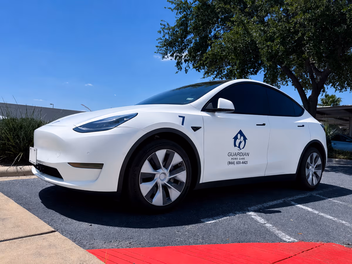 White Guardian Home Care LLC vehicle parked in a sunny outdoor lot beside a tree.