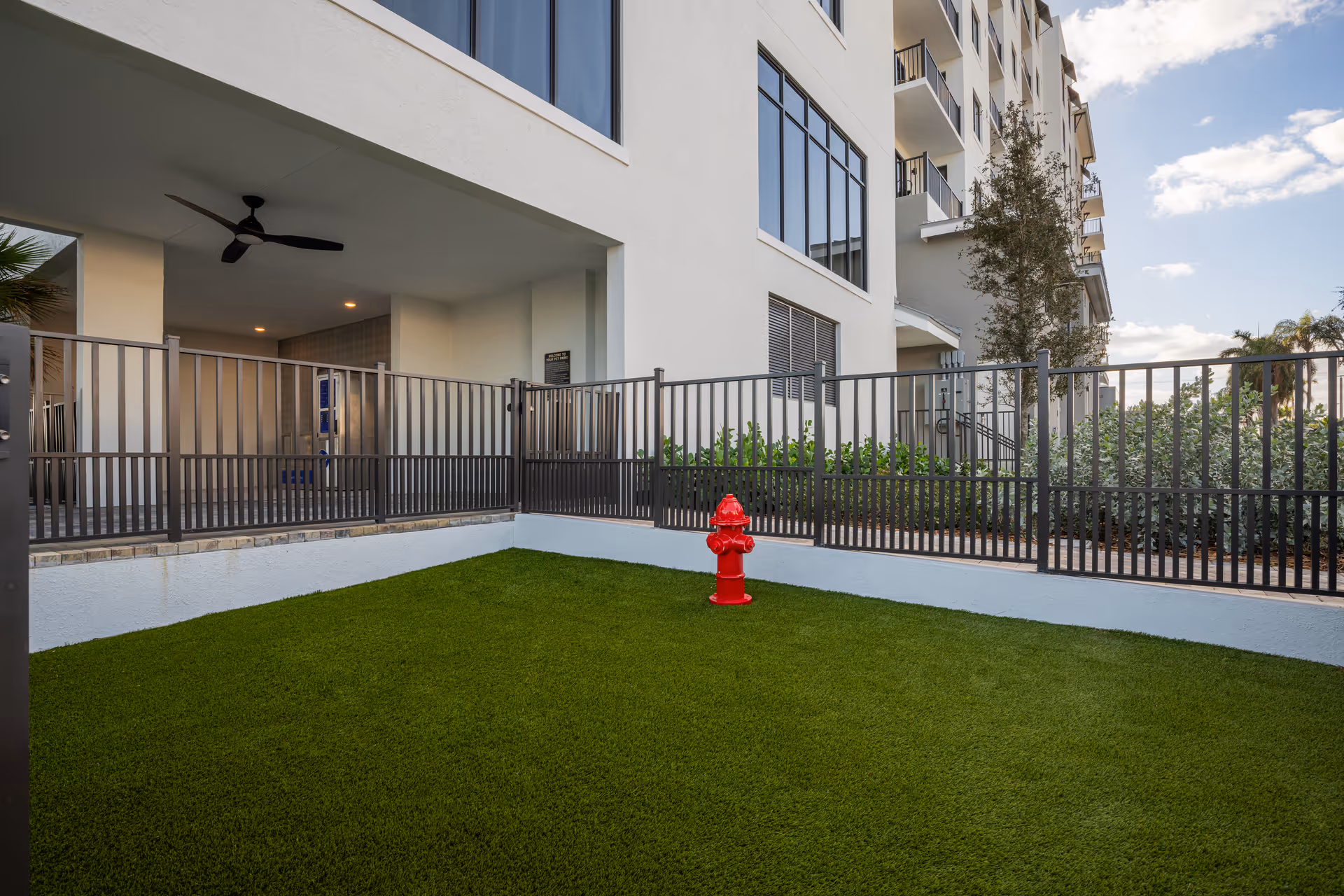 Outdoor fenced area with artificial green grass and a red fire hydrant in front of a multi-story building under a partly cloudy sky.
