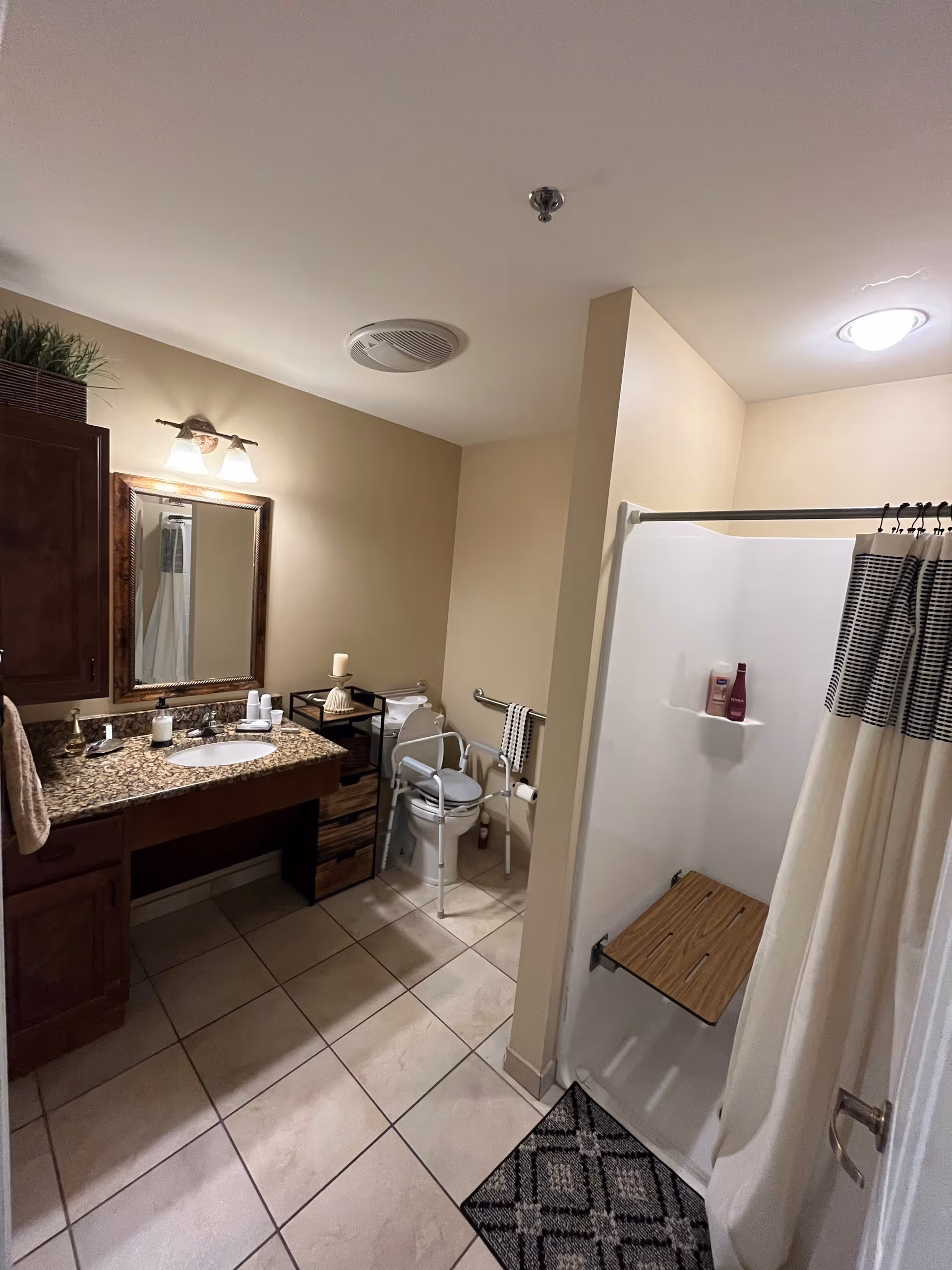A bathroom with beige walls and tiled floor featuring a granite countertop with a sink, a mirror with wooden frame, a cabinet, a toilet with safety rails and a shower area with a foldable wooden seat and a striped shower curtain.